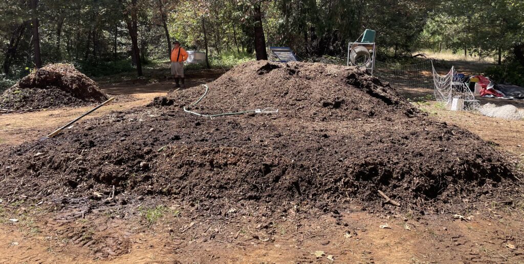 Large outdoor mound of organic material with a watering hose, trees and screening equipment nearby.