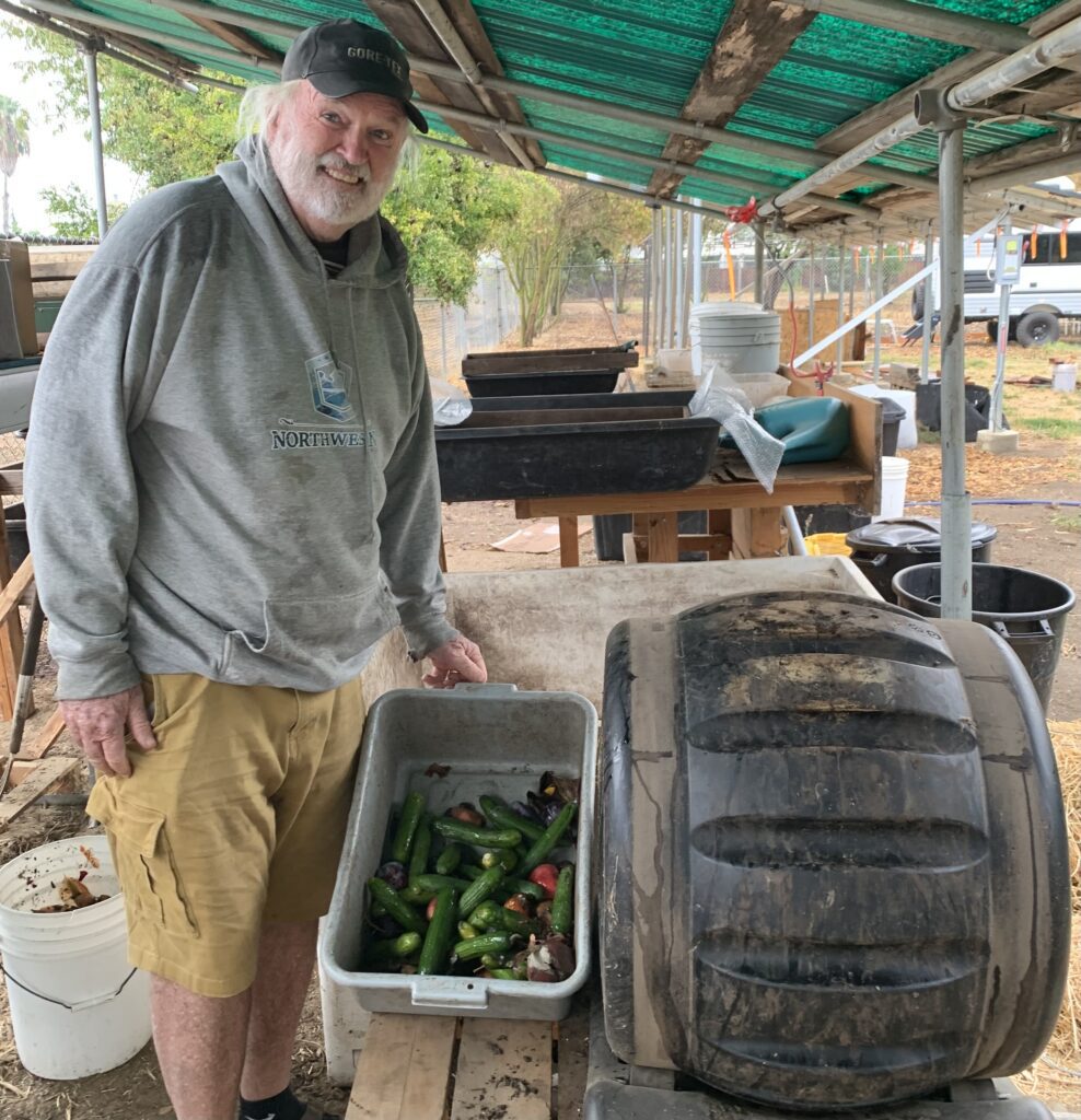 Chris Choate smiling beside a compost tumbler and a tray of fruit and vegetable scraps at Delta Worms.