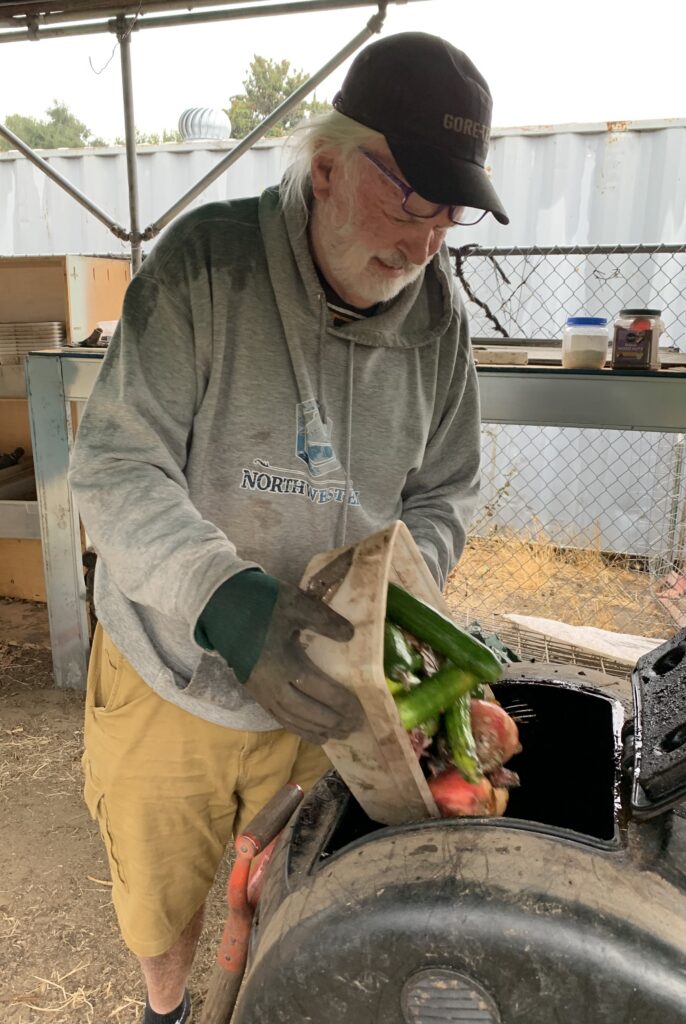 Chris Choate tipping a tray of cucumbers and fruit into an open compost tumbler.